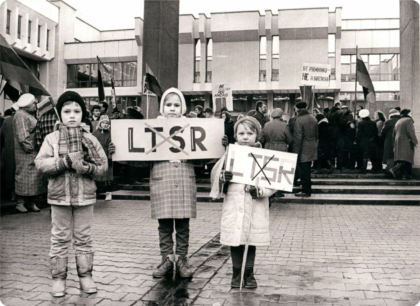 Pirmame plane trys vaikai, du iš jų laiko plakatus, kuriame nubrauktos raidės LTSR. už jų matosi Seimo rūmai ir protestuojantys žmonės.